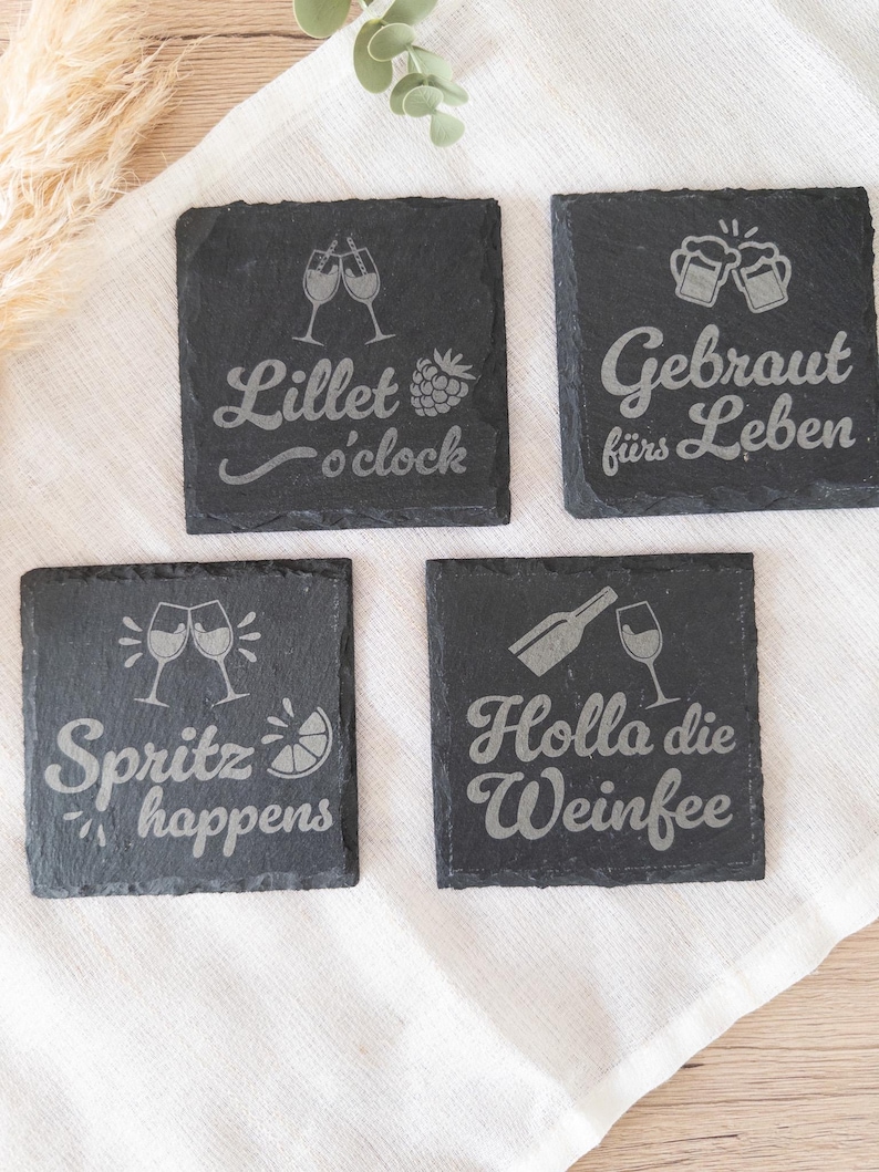 Vier quadratische Untersetzer aus schwarzem Schiefer mit gravierten Sprüchen: Lillet o’clock mit Gläsern und Himbeere, Gebraut fürs Leben mit Bierkrügen, Spritz happens mit Gläsern und Zitrone sowie Holla die Weinfee mit Flasche und Weinglas. Die Untersetzer liegen dekorativ auf einem hellen Stofftuch.