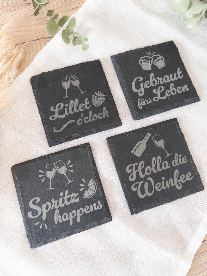 Vier quadratische Untersetzer aus schwarzem Schiefer mit gravierten Sprüchen: Lillet o’clock mit Gläsern und Himbeere, Gebraut fürs Leben mit Bierkrügen, Spritz happens mit Gläsern und Zitrone sowie Holla die Weinfee mit Flasche und Weinglas. Die Untersetzer liegen dekorativ auf einem hellen Stofftuch.