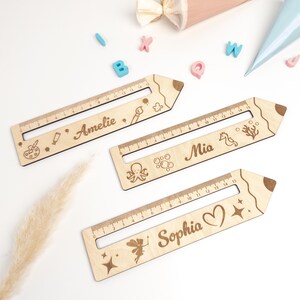 Drei personalisierte Lesehilfen aus Holz mit Lineal liegen auf hellem Hintergrund. Die Leselineale sind mit den Namen „Amelie“, „Mia“ und „Sophia“ sowie verschiedenen Motiven wie Malen, Meer und Fee graviert – ideal als Geschenk zur Einschulung oder als Inhalt für die Schultüte.