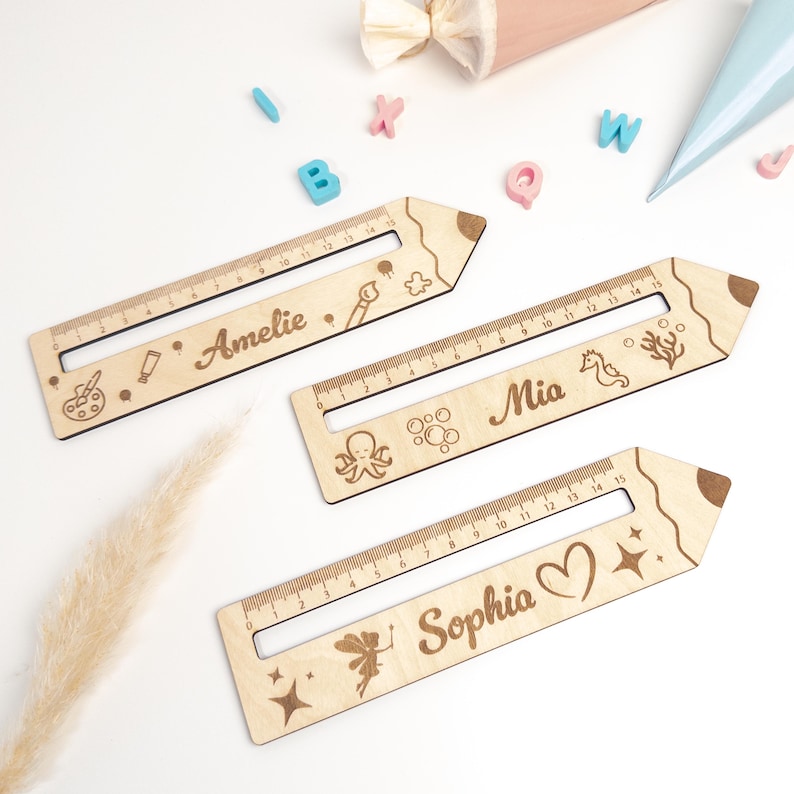 Drei personalisierte Lesehilfen aus Holz mit Lineal liegen auf hellem Hintergrund. Die Leselineale sind mit den Namen „Amelie“, „Mia“ und „Sophia“ sowie verschiedenen Motiven wie Malen, Meer und Fee graviert – ideal als Geschenk zur Einschulung oder als Inhalt für die Schultüte.