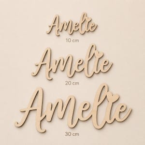 May include: Three wooden cutouts of the name "Amelie" in a cursive font. The sizes are 10 cm, 20 cm, and 30 cm. Each name has a heart detail. The wood has a natural, light brown color.