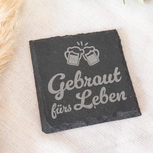 Ein quadratischer Untersetzer aus schwarzem Schiefer mit graviertem Spruch:  Gebraut fürs Leben mit Bierkrügen. Der Untersetzer liegt dekorativ auf einem hellen Stofftuch.