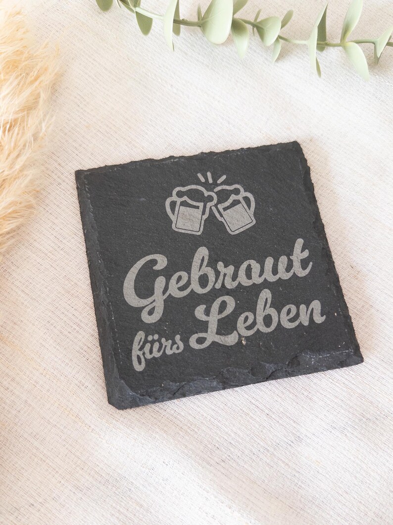 Ein quadratischer Untersetzer aus schwarzem Schiefer mit graviertem Spruch:  Gebraut fürs Leben mit Bierkrügen. Der Untersetzer liegt dekorativ auf einem hellen Stofftuch.