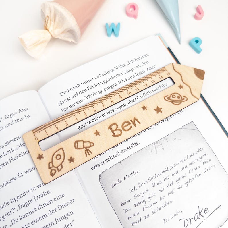 Personalisierte Lesehilfe aus Holz mit Lineal und Weltraum-Motiv liegt auf einem geöffneten Kinderbuch. Das Leselineal ist mit dem Namen „Ben“, Rakete, Planeten und Sternen graviert und eignet sich als Geschenk zur Einschulung oder als Inhalt für die Schultüte.