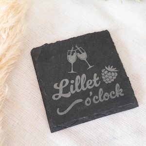 Ein quadratischer Untersetzer aus schwarzem Schiefer mit graviertem Spruch:  Lillet o’clock mit Gläsern und Himbeere. Der Untersetzer liegt dekorativ auf einem hellen Stofftuch.
