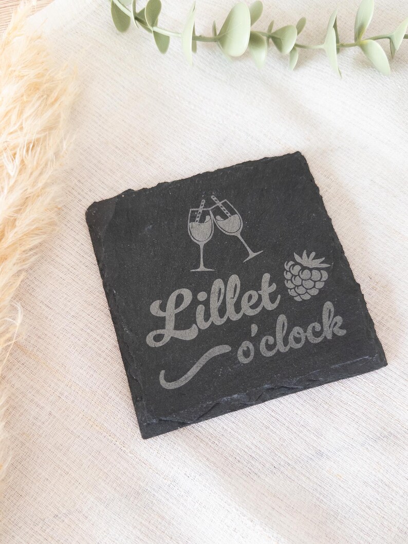Ein quadratischer Untersetzer aus schwarzem Schiefer mit graviertem Spruch:  Lillet o’clock mit Gläsern und Himbeere. Der Untersetzer liegt dekorativ auf einem hellen Stofftuch.