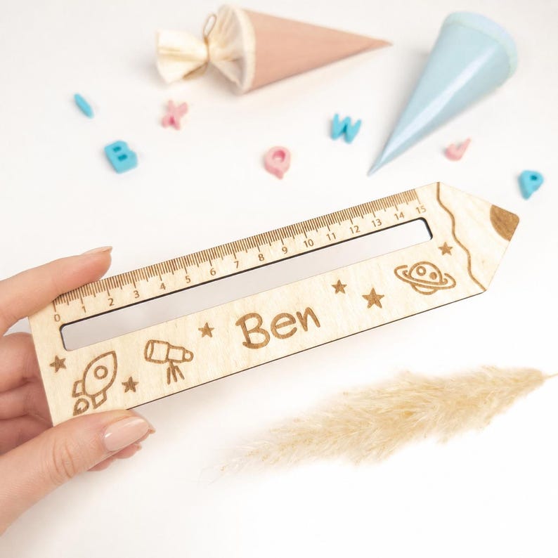 Personalisierte Lesehilfe aus Holz mit Lineal wird in der Hand gehalten. Das Leselineal ist mit dem Namen „Ben“ sowie Rakete, Teleskop, Sternen und Planet graviert – ideal als Geschenk zur Einschulung und als Inhalt für die Schultüte.