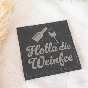 Ein quadratischer Untersetzer aus schwarzem Schiefer mit graviertem Spruch:  Holla die Weinfee mit Flasche und Weinglas. Der Untersetzer liegt dekorativ auf einem hellen Stofftuch.