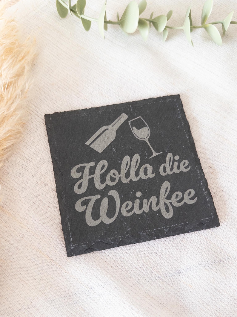 Ein quadratischer Untersetzer aus schwarzem Schiefer mit graviertem Spruch:  Holla die Weinfee mit Flasche und Weinglas. Der Untersetzer liegt dekorativ auf einem hellen Stofftuch.