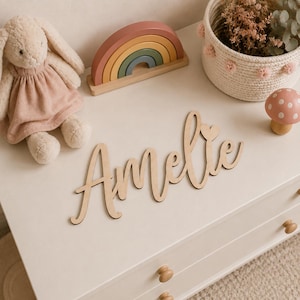 May include: A wooden name cutout that spells "Amelie" in a cursive font, with a small heart above the "i". The name is displayed on a white dresser, alongside a plush bunny, a wooden rainbow, and a mushroom figurine.