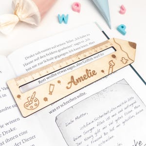 Personalisierte Lesehilfe aus Holz mit Lineal liegt auf einem geöffneten Kinderbuch. Das Leselineal ist mit dem Namen „Amelie“ sowie Mal-Motiven wie Farbpalette, Stift, Pinsel und Klecks graviert – ideal als Geschenk zur Einschulung und als Inhalt für die Schultüte.