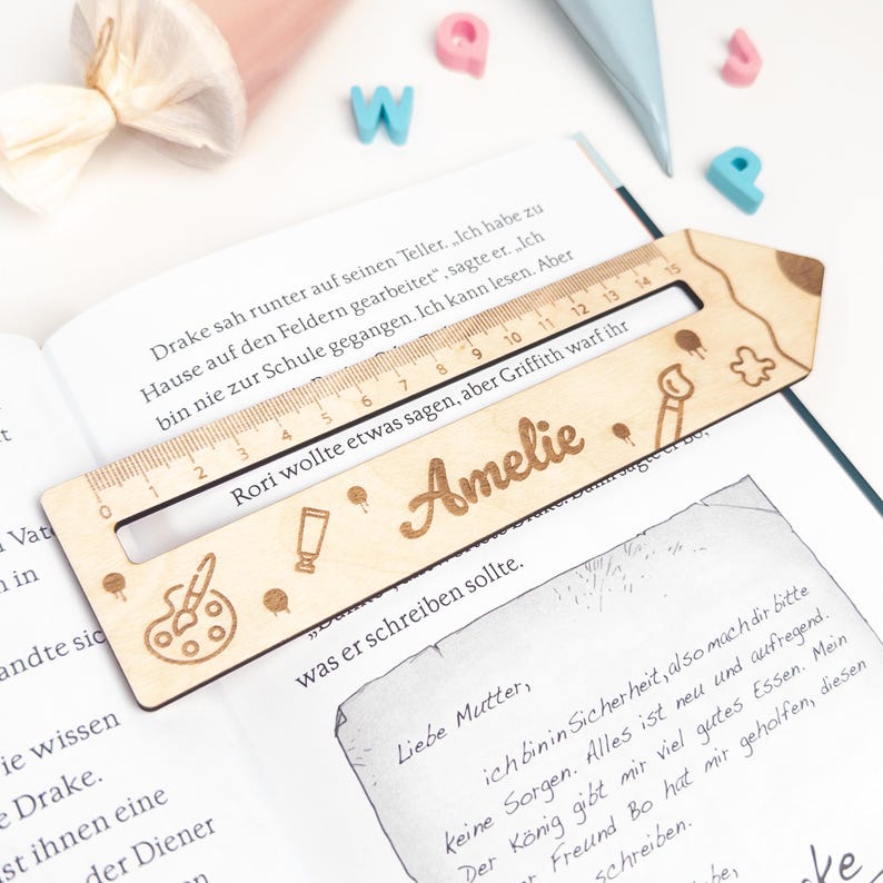 Personalisierte Lesehilfe aus Holz mit Lineal liegt auf einem geöffneten Kinderbuch. Das Leselineal ist mit dem Namen „Amelie“ sowie Mal-Motiven wie Farbpalette, Stift, Pinsel und Klecks graviert – ideal als Geschenk zur Einschulung und als Inhalt für die Schultüte.