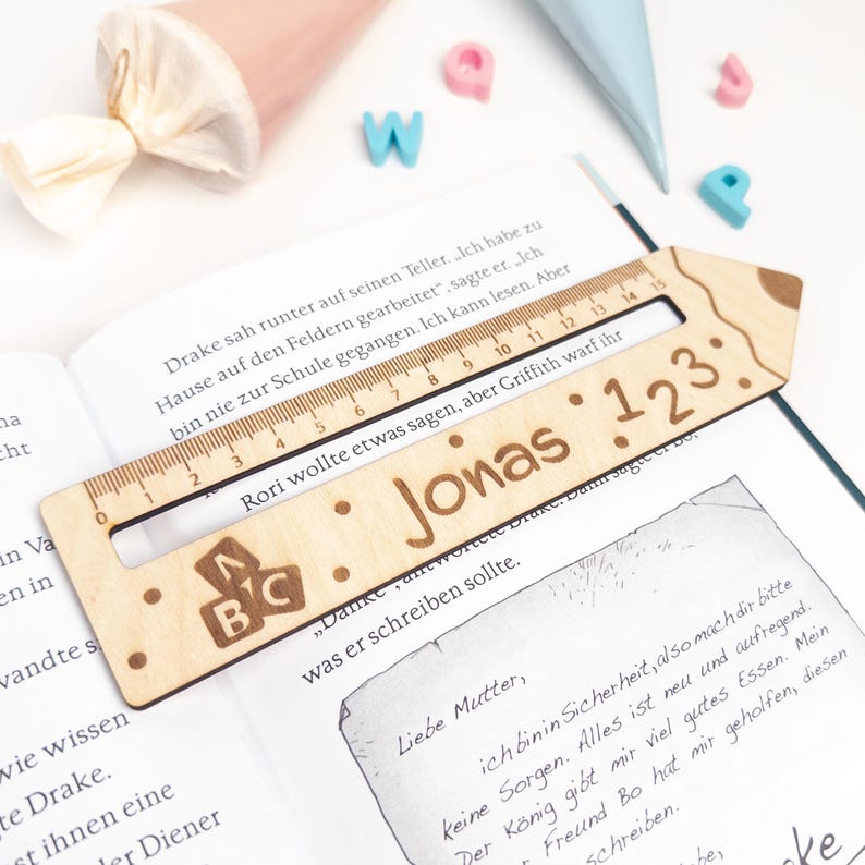 Personalisierte Lesehilfe aus Holz mit Lineal liegt auf einem geöffneten Kinderbuch. Das Leselineal ist mit dem Namen „Jonas“, ABC-Motiv und „1 2 3“ graviert – eine praktische Geschenkidee zur Einschulung und ideal als Inhalt für die Schultüte.
