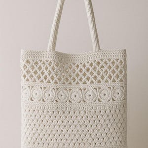 May include: Cream-colored crochet tote bag with two handles. The bag features a detailed pattern with diamond and floral designs. The bag is made of a soft, textured yarn, perfect for carrying everyday essentials.