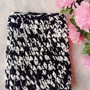 Op de afbeelding: Een vierkant, handgehaakt item in een zwart-wit patroon. Het item is gemaakt van dik garen en heeft een textuur. Roze bloemen staan op de achtergrond.