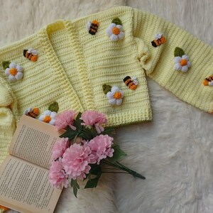 May include: A light yellow crochet cardigan adorned with 3D bee and daisy appliqués. The sleeves have orange stripes at the cuffs. A book and pink flowers are in the foreground, all on a fluffy white surface.