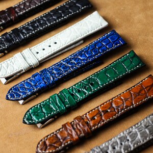 Peut inclure: Une collection de bracelets de montre de différentes couleurs, dont blanc, bleu, vert et marron. Chaque bracelet présente un motif texturé en forme de crocodile avec des coutures blanches contrastantes et des boucles argentées. Les bracelets sont disposés sur une surface de ton neutre.