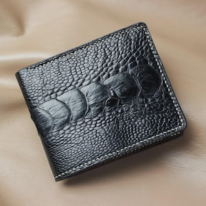 May include: A black bifold wallet featuring a textured ostrich leg skin pattern with large, prominent scales running across the center. The wallet is constructed with visible white stitching along the edges, creating a distinct contrast against the dark surface.