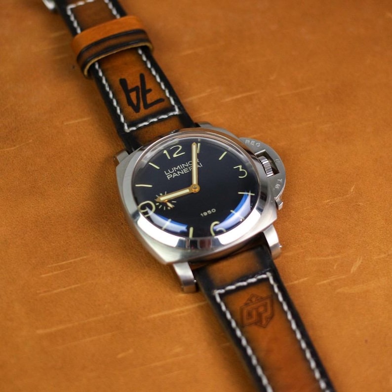 May include: A stainless steel wristwatch with a black face and gold hands, featuring the text "Luminor Panerai 1950". The watch has a brown leather band with white stitching and the number "74" printed on it. The watch is displayed on a brown surface.