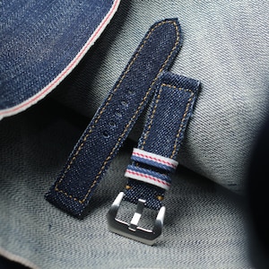Può includere: Un cinturino per orologio in denim blu scuro con cuciture color oro lungo i bordi. Il cinturino include due passanti in tessuto con dettagli a righe rosse e bianche e una fibbia in metallo color argento lucido per il fissaggio all'orologio.