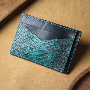 May include: A rectangular card holder featuring a combination of smooth black leather and a textured teal and black snakeskin-patterned leather. The accessory is constructed with visible teal stitching along the edges and a V-shaped pocket opening.