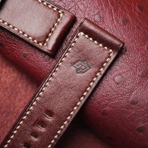 May include: Close-up of a brown leather watch strap with cream-colored stitching. The strap has a stamped logo and several adjustment holes. The background is a textured, reddish-brown leather surface.