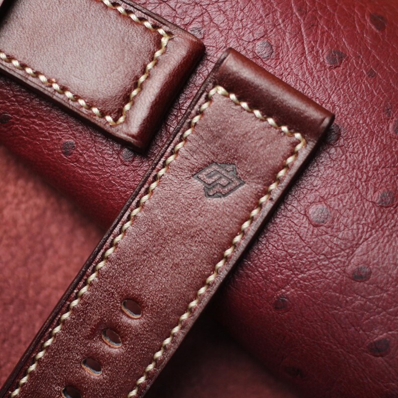 May include: Close-up of a brown leather watch strap with cream-colored stitching. The strap has a stamped logo and several adjustment holes. The background is a textured, reddish-brown leather surface.