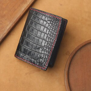 May include: A black bi-fold wallet featuring a textured crocodile skin pattern on the exterior. The wallet is accented with vibrant red stitching along the edges, providing a distinct contrast against the dark, glossy surface of the leather material.