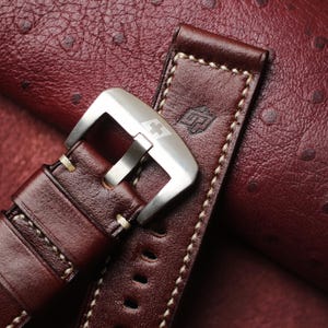 May include: Close-up of a brown leather watch strap with cream stitching and a silver buckle. The strap features a stamped logo and is set against a textured, reddish-brown background.