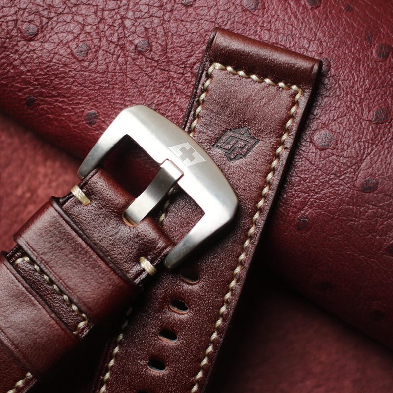 May include: Close-up of a brown leather watch strap with cream stitching and a silver buckle. The strap features a stamped logo and is set against a textured, reddish-brown background.