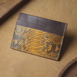 May include: A rectangular card holder featuring a smooth dark brown leather top section and a textured yellow and black snakeskin pattern on the lower portion, finished with visible yellow stitching along the edges of the leather construction.