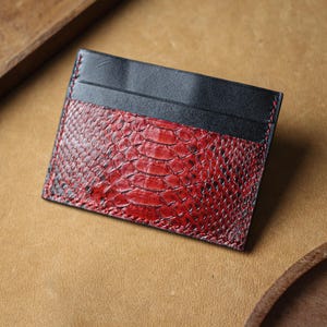 May include: A rectangular card holder featuring a smooth black leather top section and a textured red snakeskin pattern on the lower portion. The edges are finished with visible red stitching, creating a contrast between the two distinct materials.