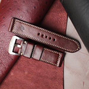 May include: A close-up of a brown leather watch strap with white stitching. The strap has a silver buckle and a stamped logo. The leather has a rich, dark color and a textured appearance. The watch strap is a replacement part.