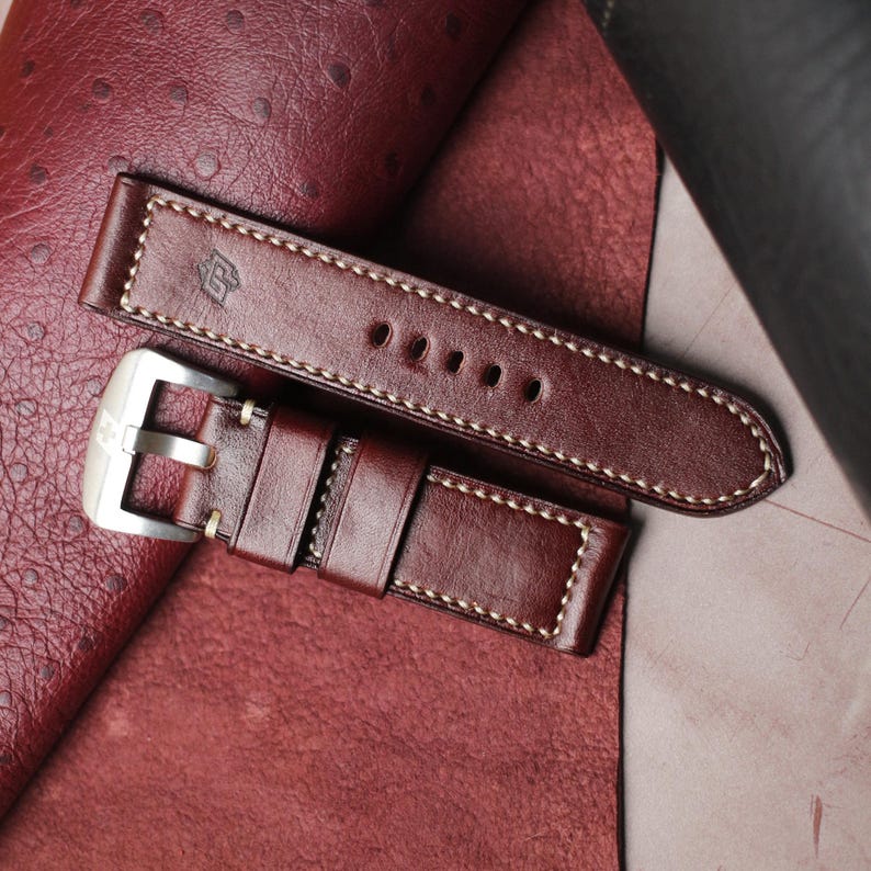 May include: A close-up of a brown leather watch strap with white stitching. The strap has a silver buckle and a stamped logo. The leather has a rich, dark color and a textured appearance. The watch strap is a replacement part.