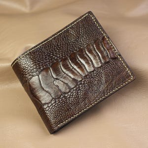 May include: A dark brown bifold wallet featuring a textured ostrich leg leather pattern. The exterior displays a distinct combination of small, pebbled scales and a central vertical row of larger, rectangular scales, finished with light stitching.