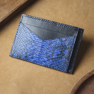 May include: A rectangular card holder featuring a combination of smooth black leather and a textured blue and black snakeskin pattern. The design includes a V-shaped top pocket edge with visible stitching along the perimeter of the accessory.