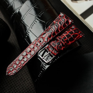 Puede incluir: Un par de correas de reloj con un patrón de cocodrilo rojo y negro. Las correas tienen un acabado brillante y una hebilla plateada. Las correas de reloj son de cuero.