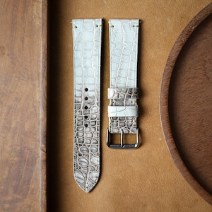 Puede incluir: Dos correas de reloj gris claro con un patrón de cocodrilo. Las correas son de cuero y tienen una hebilla plateada. Las correas se muestran sobre una superficie marrón.