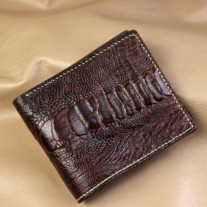 Peut inclure: Un portefeuille bifold marron foncé présentant un motif en cuir d'autruche texturé avec une rangée d'écailles plus grandes et surélevées en diagonale sur le devant. Le portefeuille est fini avec des coutures claires visibles sur les bords.