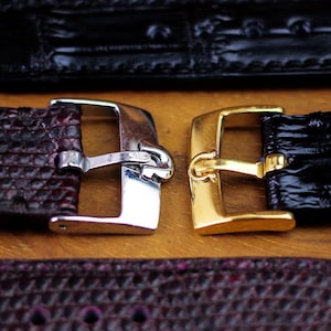 Pode incluir: Close-up de duas pulseiras de relógio com fivelas de metal. Uma pulseira é bordô escuro com fivela prateada, e a outra é preta com fivela dourada. As pulseiras têm um design texturizado.