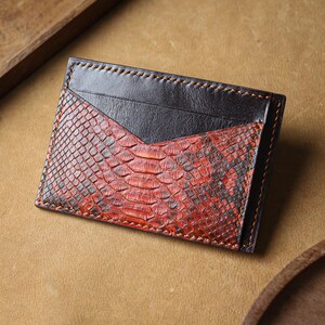 May include: A rectangular card holder featuring a combination of smooth black leather and a textured red and black snakeskin pattern. The accessory is constructed with visible orange stitching along the edges and a V-shaped pocket design.