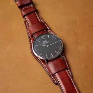 May include: A wristwatch with a black watch face and silver-colored hands and casing. The watch is attached to a wide, brown leather band with white stitching. The watch face has the text "DW Daniel Wellington".