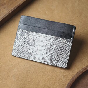 May include: A rectangular card holder featuring a combination of smooth black leather at the top and a textured, silver and white snakeskin-patterned leather on the lower section, finished with visible white stitching along the edges.