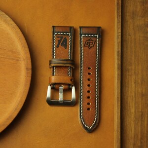 May include: Two brown leather watch straps with white stitching. One strap features the number "74" in black, and the other has a logo. The straps have a silver buckle and are displayed on a brown surface.
