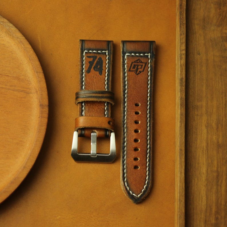May include: Two brown leather watch straps with white stitching. One strap features the number "74" in black, and the other has a logo. The straps have a silver buckle and are displayed on a brown surface.