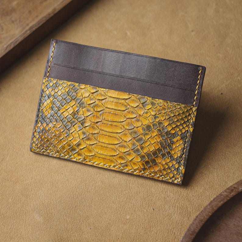 May include: A rectangular card holder featuring a combination of smooth dark brown leather on the top section and a textured yellow and black snakeskin pattern on the bottom section, finished with visible yellow stitching along the edges.