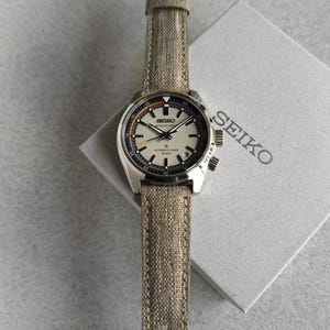 Può includere: Un orologio Seiko Speedtimer argentato con cinturino in tela beige. Il quadrante dell'orologio è bianco sporco con dettagli neri e arancioni. L'orologio è presentato su una scatola bianca con il logo Seiko.