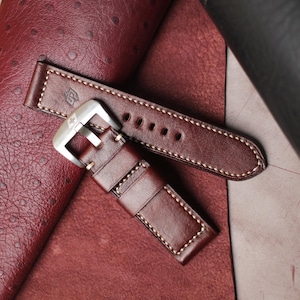 May include: A brown leather watch strap with white stitching and a silver buckle. The strap has a classic design with a logo embossed on the leather. The watch band is shown on a textured, reddish-brown surface.