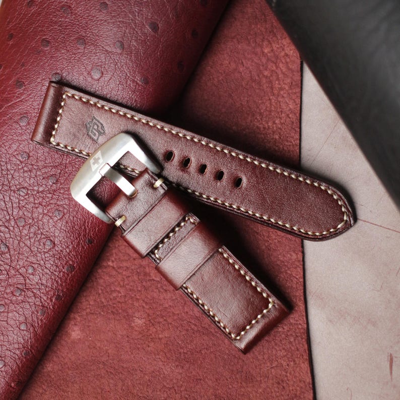 May include: A brown leather watch strap with white stitching and a silver buckle. The strap has a classic design with a logo embossed on the leather. The watch band is shown on a textured, reddish-brown surface.