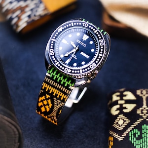 Puede incluir: Un reloj Seiko con esfera negra y correa estampada en verde, amarillo y negro. El reloj tiene un bisel plateado con marcas blancas. La correa tiene un diseño geométrico. El reloj se exhibe sobre un soporte.